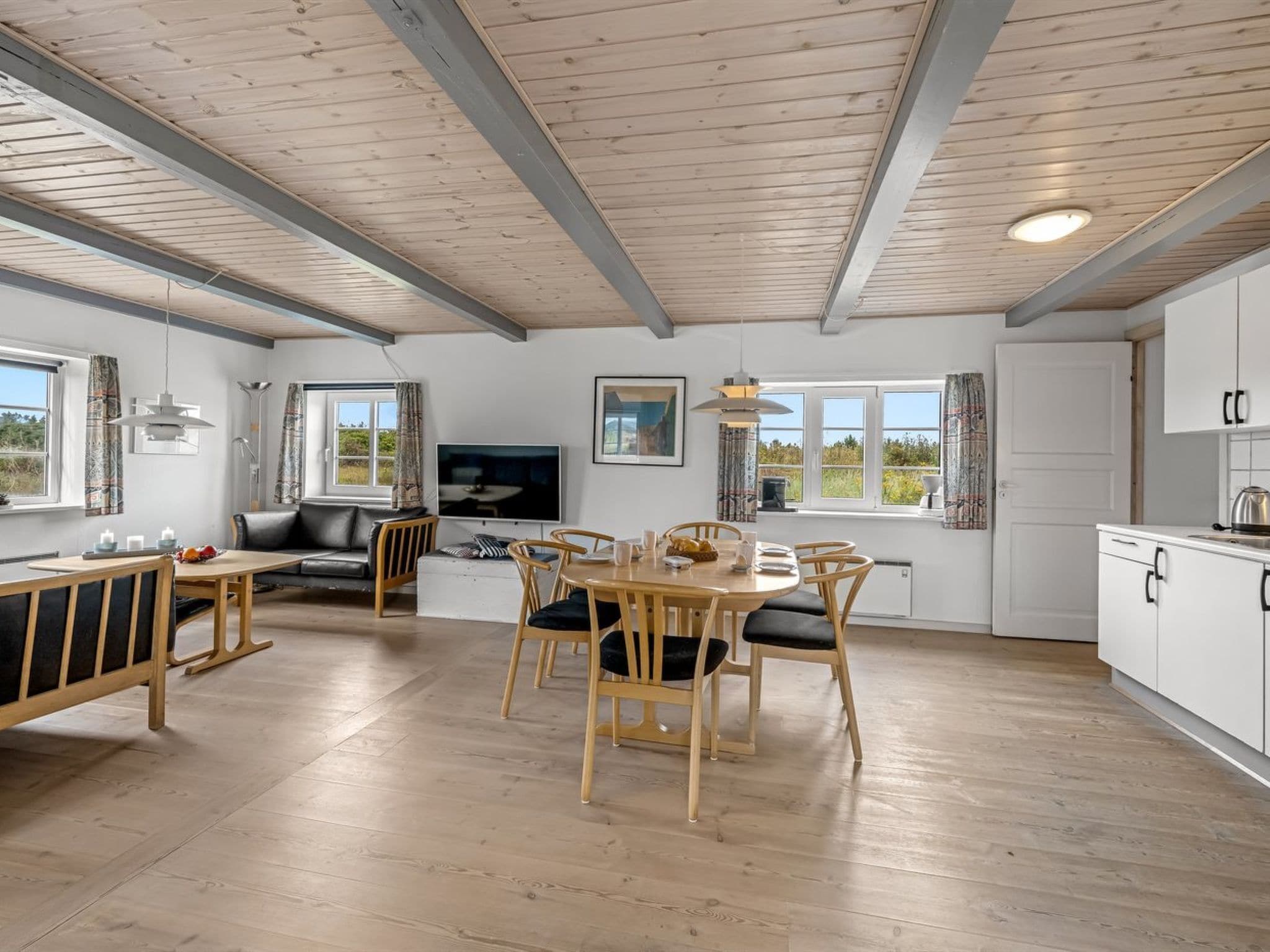 Een moderne keuken met witte kastjes, een eethoek met houten stoelen en een tafel, en een zithoek met een bank en televisie.