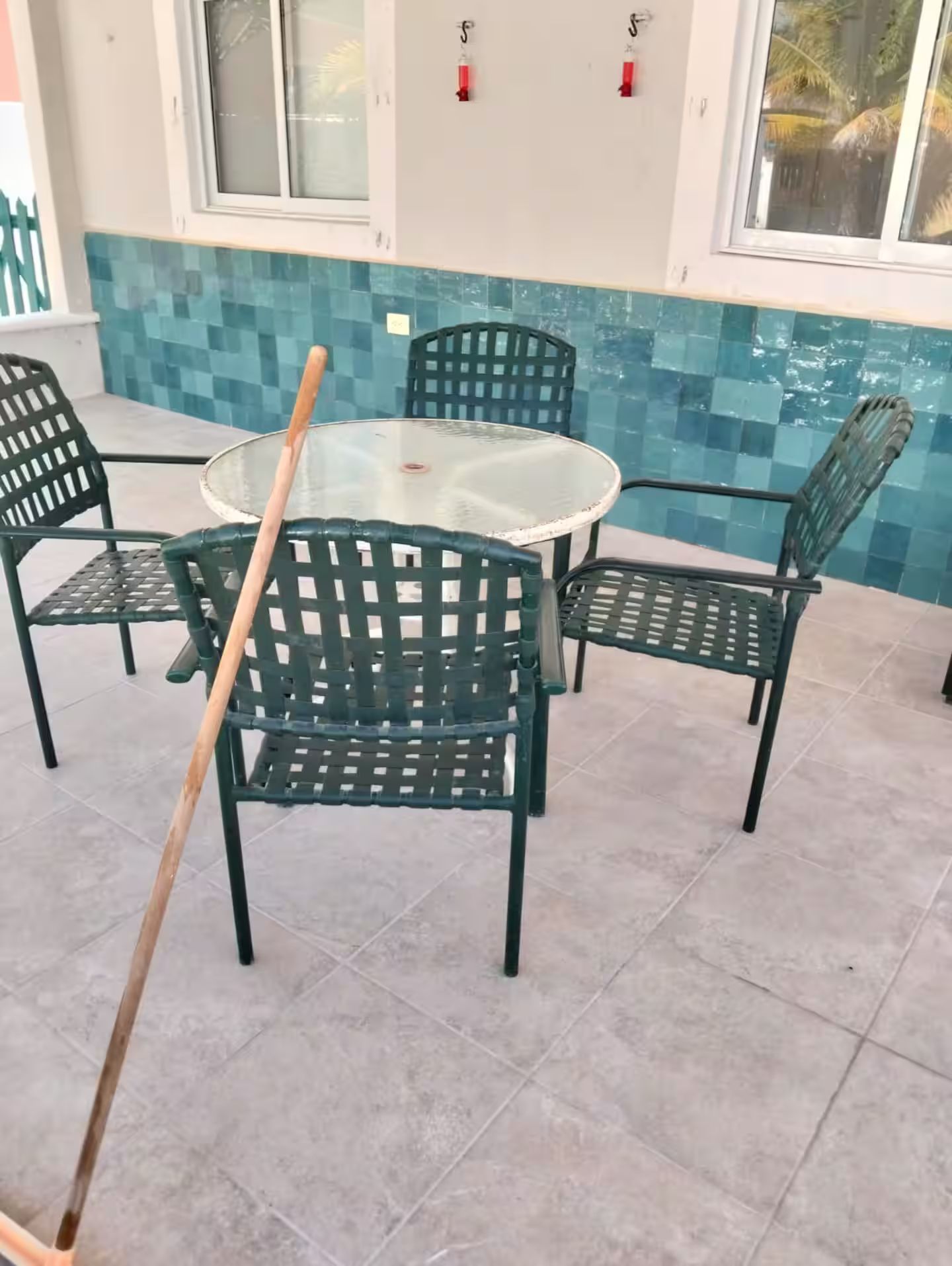Eine Terrassenfläche mit einem runden Tisch und vier grünen Stühlen, ein Besen, der an einem Stuhl angelehnt ist, und eine mit blauem Mosaikmuster versehene geflieste Wand.
