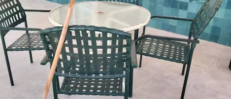 Eine Terrassenfläche mit einem runden Tisch und vier grünen Stühlen, ein Besen, der an einem Stuhl angelehnt ist, und eine mit blauem Mosaikmuster versehene geflieste Wand.