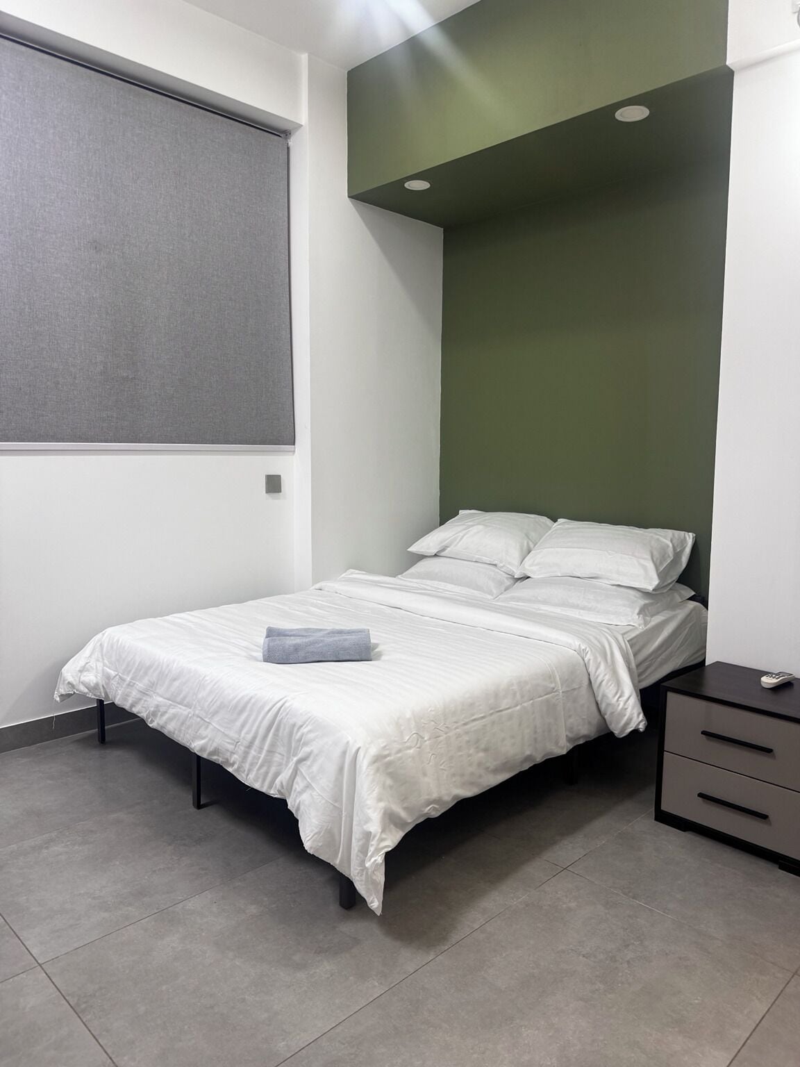Un dormitorio con cama, mesita de noche, ventana con persianas y una pared de acento verde.
