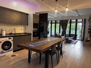 A modern kitchen with built-in appliances, a dining area with a wooden table, and a view of the outdoors through large windows.