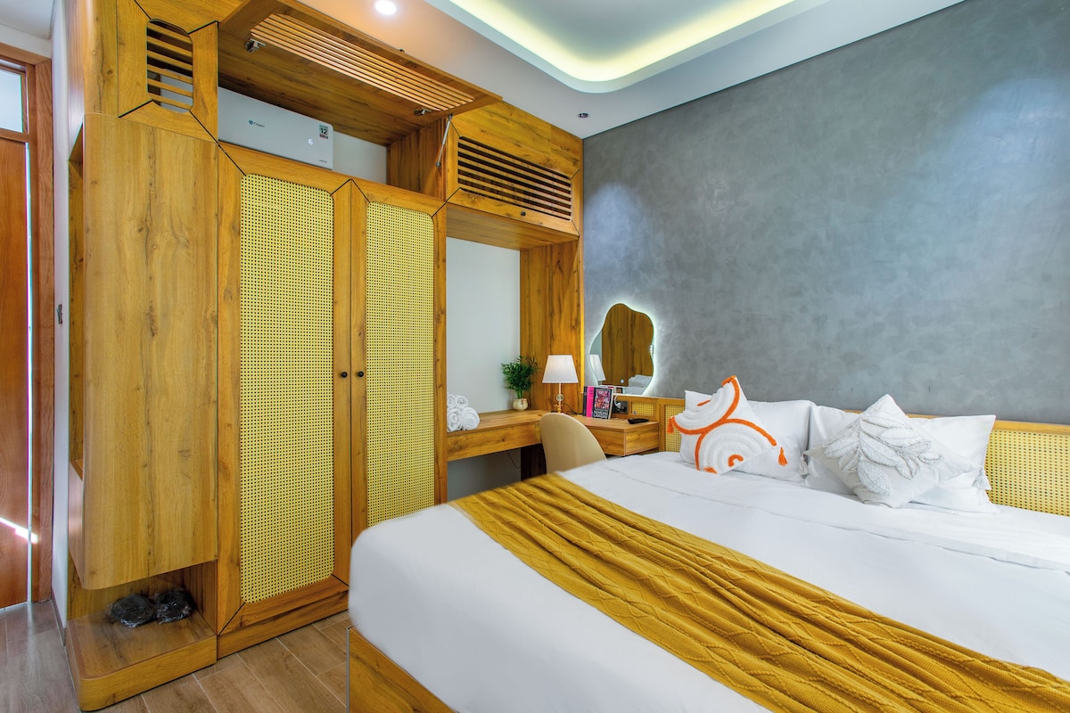 A hotel room with a bed, a wooden wardrobe, a desk with a lamp, and a large mirror.
