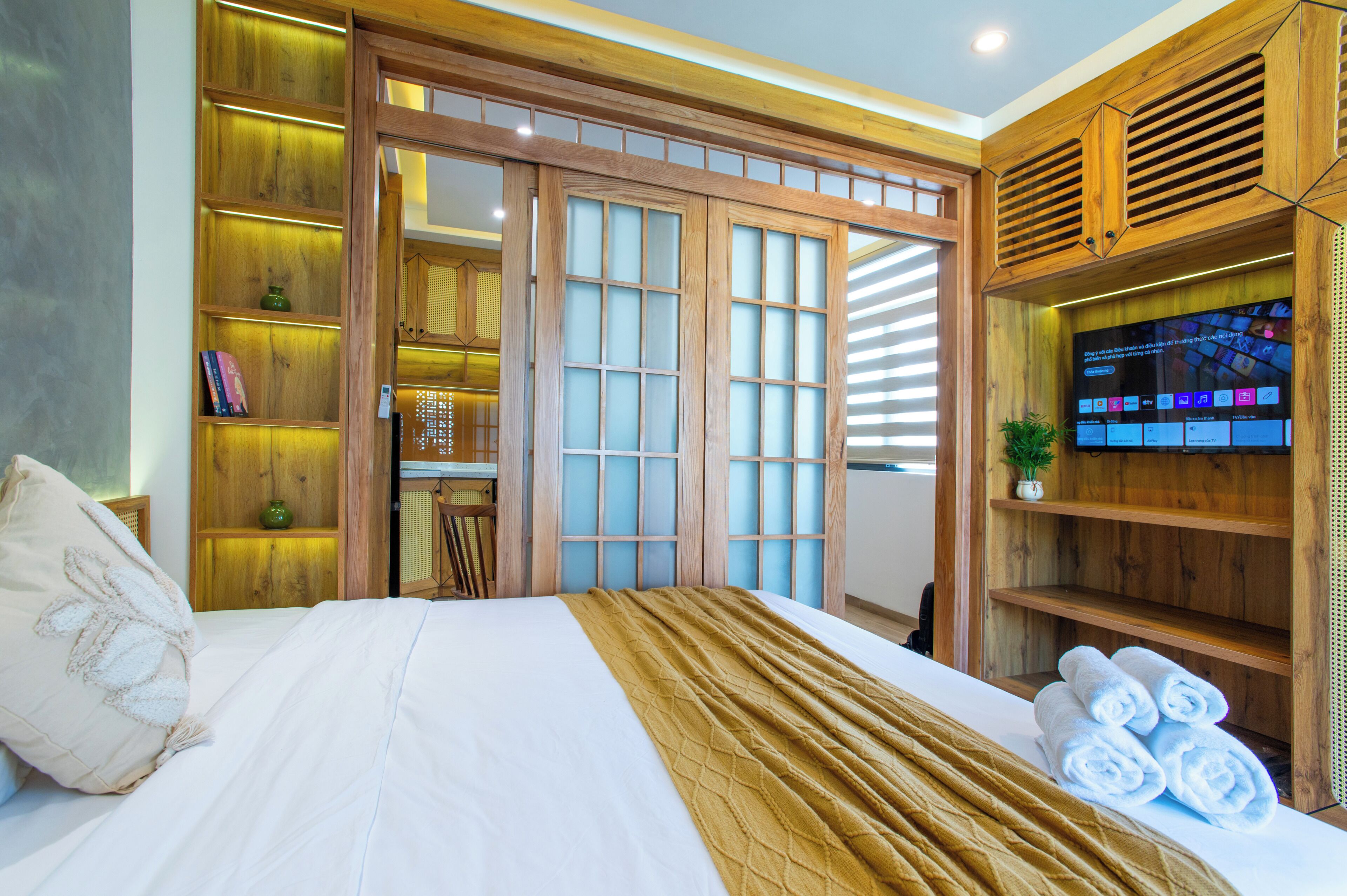 A hotel room with a bed, a TV, a wooden wardrobe, and a shelf with books and a plant.