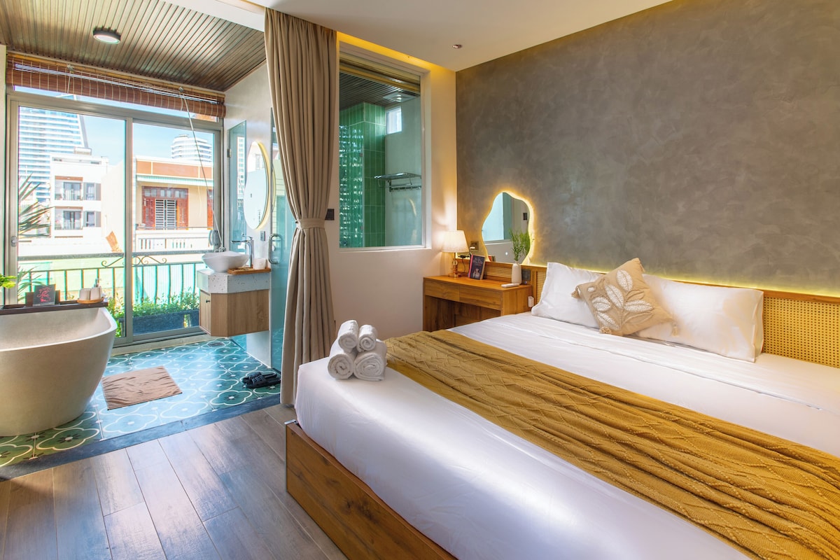 A hotel room with a large bed, a bedside table with a lamp, a mirror, and a view of the outside balcony.