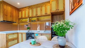 A modern kitchen with wooden cabinets, a marble countertop, a sink, and a dining area with a vase of flowers. - Lavi Residence (Da Nang)