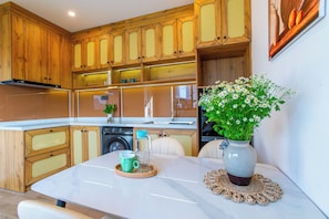 A modern kitchen with wooden cabinets, a marble countertop, a sink, and a dining area with a vase of flowers. - Lavi Residence (Da Nang)