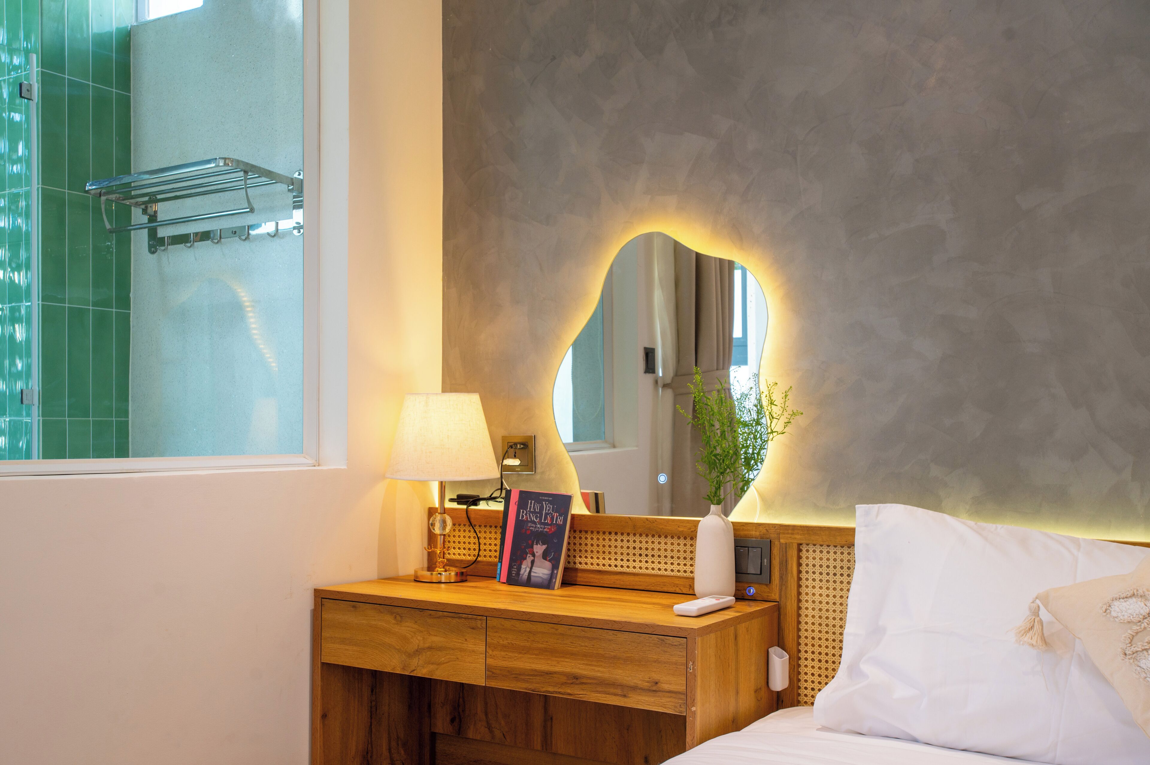 A hotel room with a wooden bedside table, a mirror with a light, a bedside lamp, a book, a small plant, and a white bed.