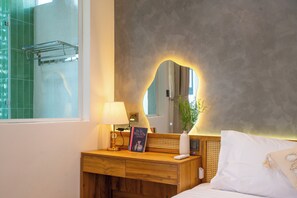 Habitación de hotel con una mesita de noche de madera, un espejo con luz, una lámpara de noche, un libro, una planta pequeña y una cama blanca.