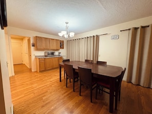 A dining area with a wooden table and chairs, a kitchen with wooden cabinets, a microwave, and a chandelier.