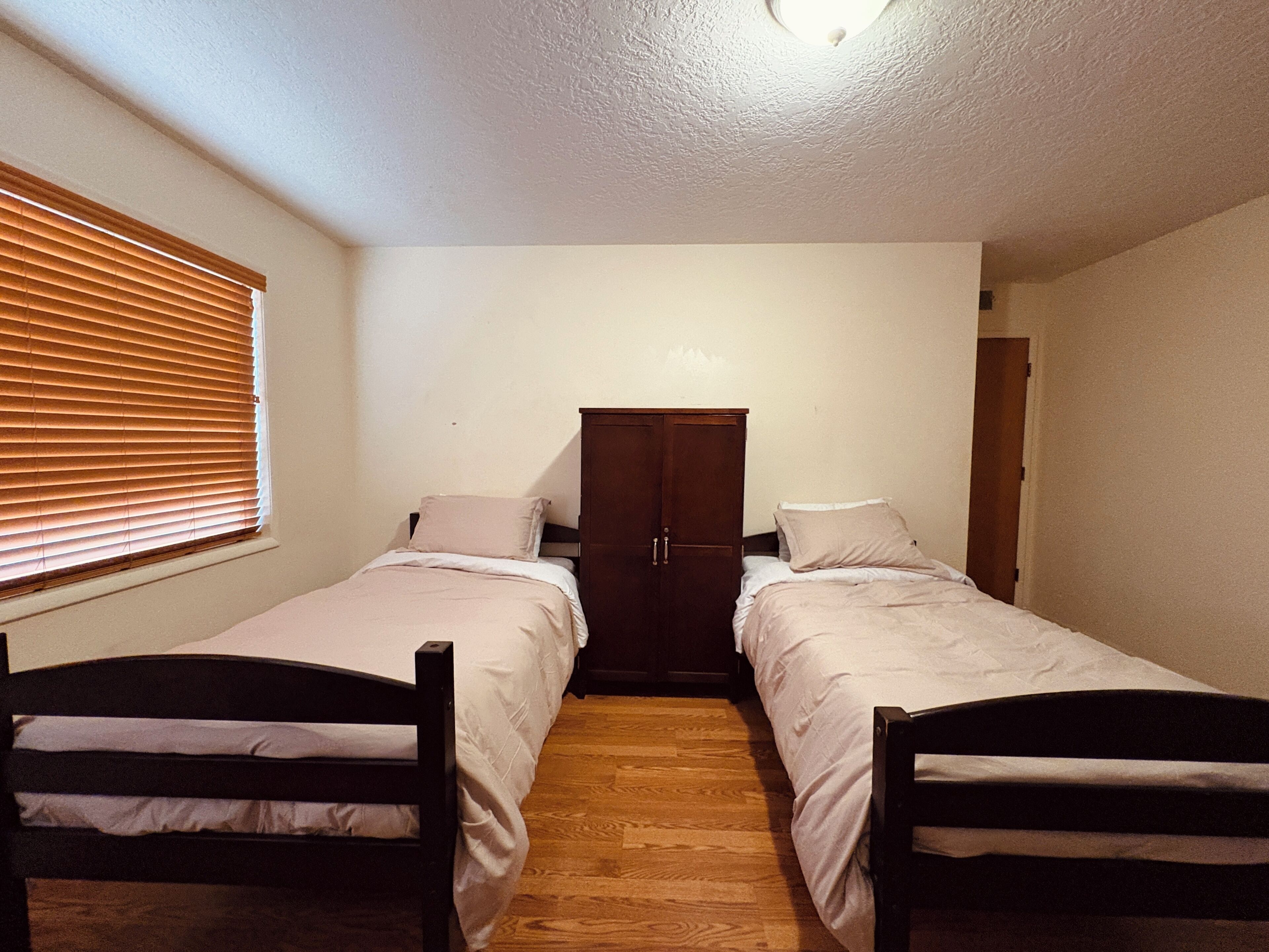 A small, single-room accommodation with two single beds, a wooden wardrobe, and a window with blinds.