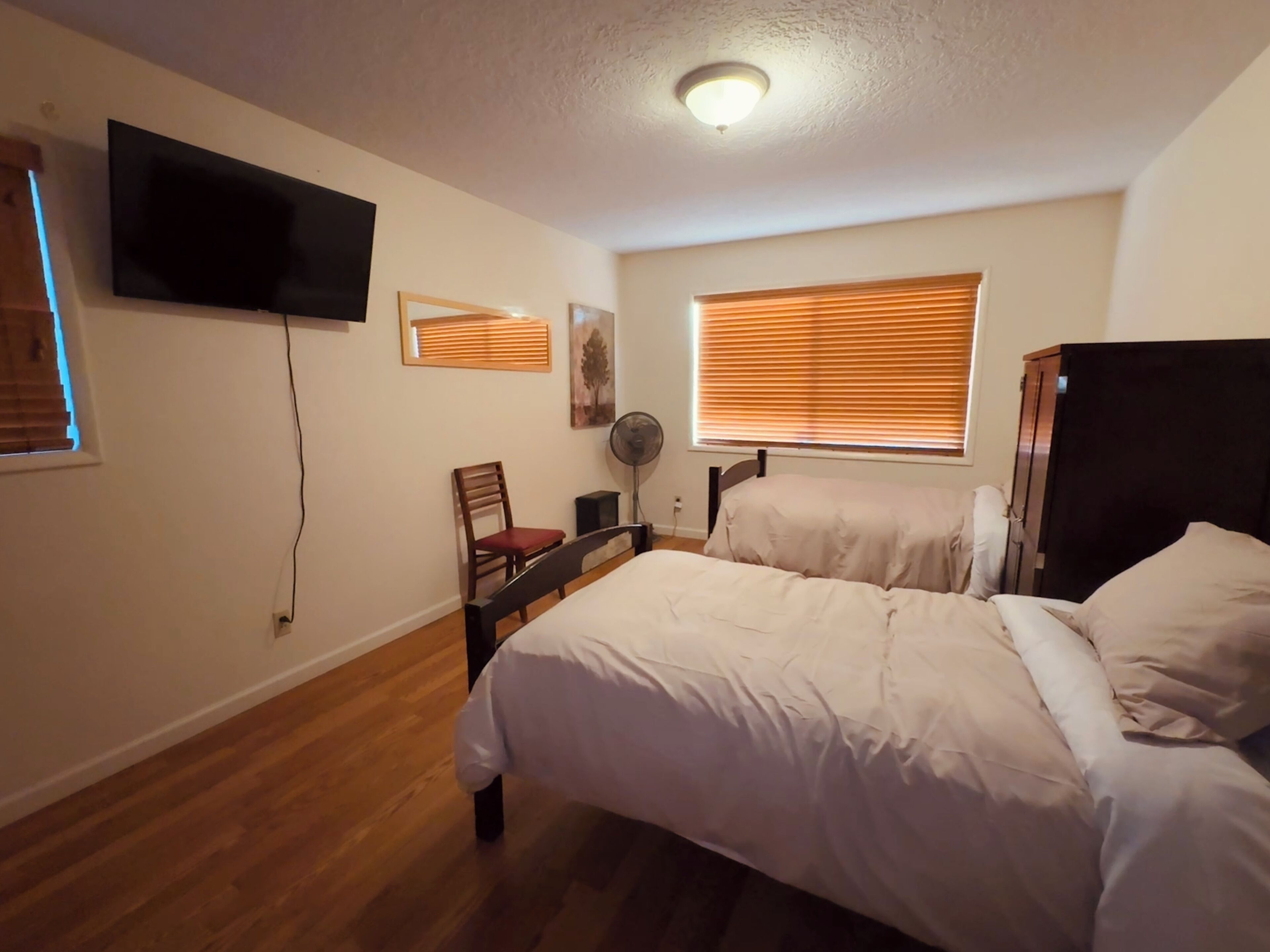 A bedroom with two beds, a TV mounted on the wall, a chair, a fan, and a window with blinds.