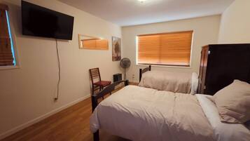 A bedroom with two beds, a TV mounted on the wall, a chair, a fan, and a window with blinds.