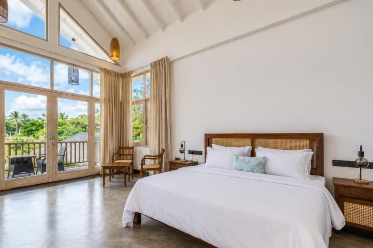 A spacious bedroom with a large bed, a wooden headboard, and a view of a lush garden through large windows.