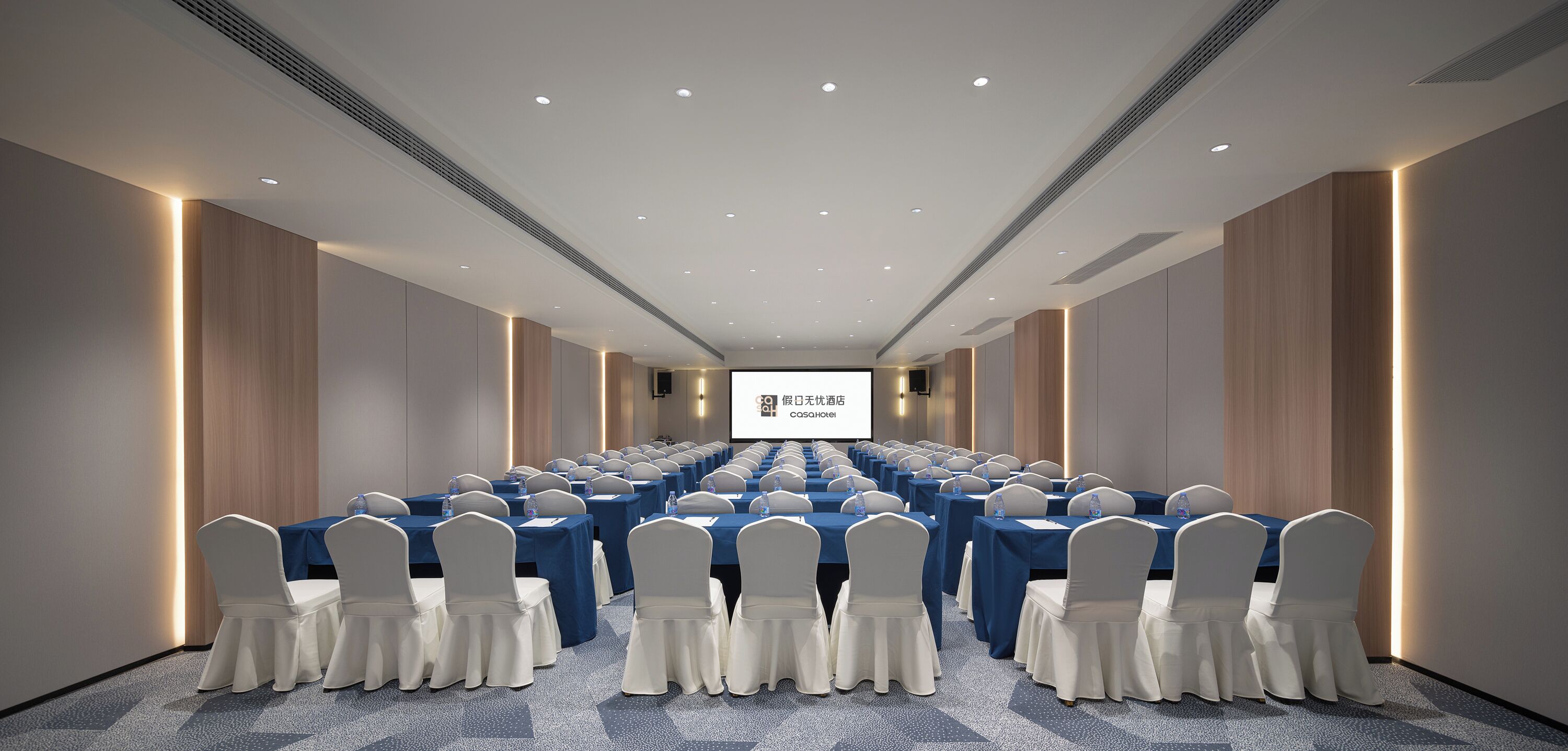 Un salón de conferencias con filas de mesas y sillas, una pantalla de proyector y un sistema de sonido.