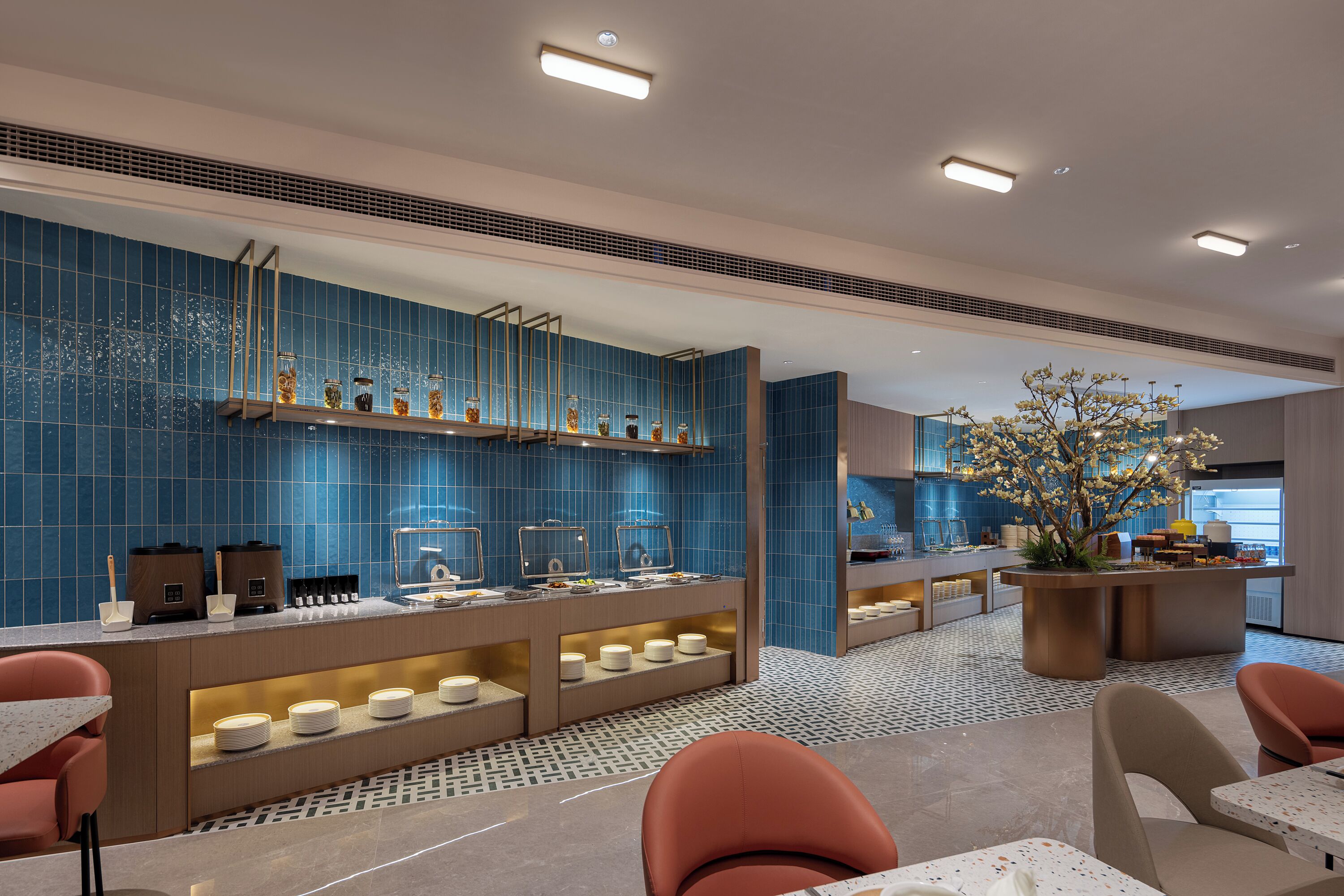 Un moderno vestíbulo de hotel con una pared de azulejos azules, una zona de buffet con mostradores de comida y diferentes disposiciones de asientos.