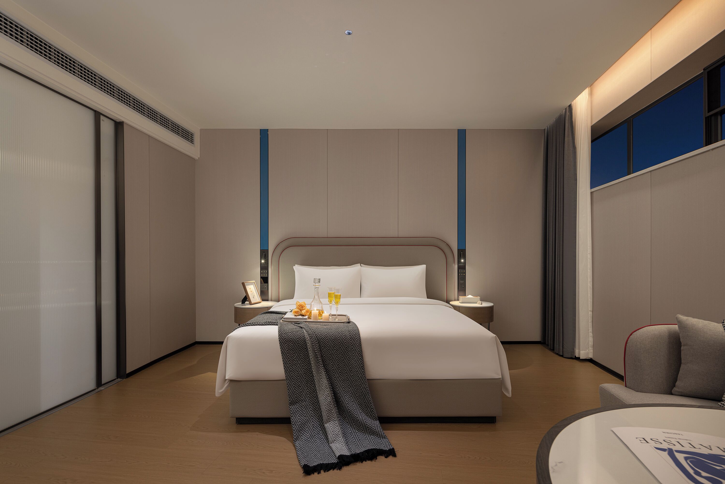 Quarto de hotel moderno com uma cama grande, mesas de cabeceira, uma poltrona e vista para o exterior.