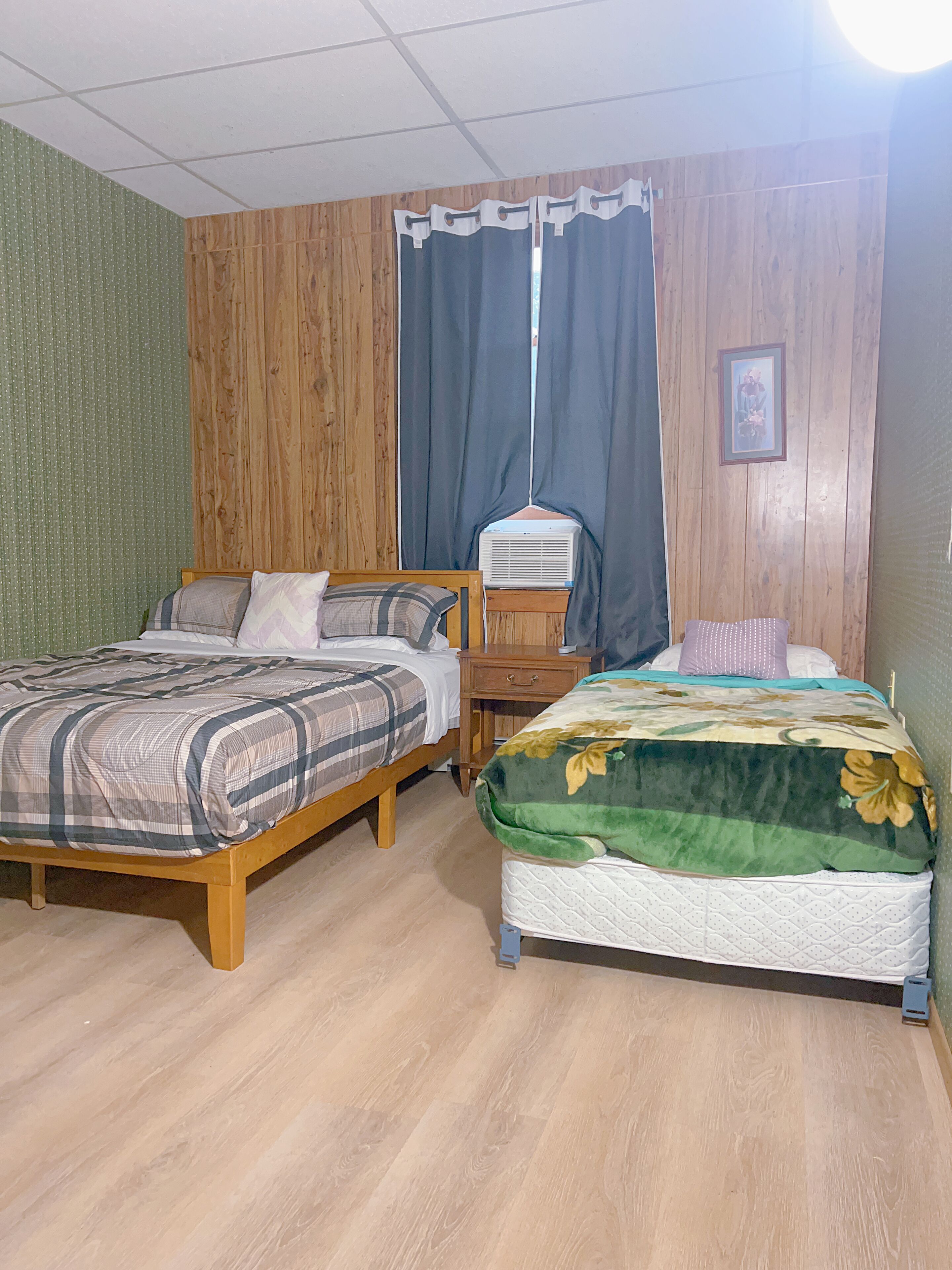 Une chambre avec deux lits en bois, une table de chevet en bois, un climatiseur fixé au mur et une fenêtre avec des rideaux.