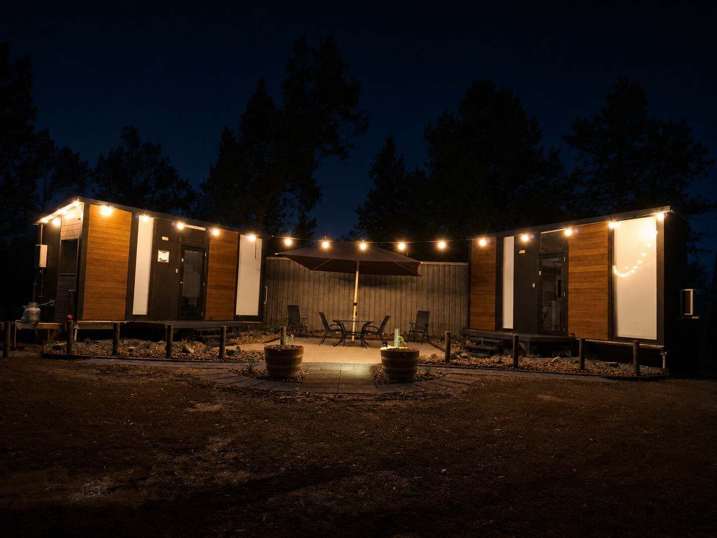 A modern outdoor seating area with a fire pit, surrounded by wooden deck and chairs, under string lights.