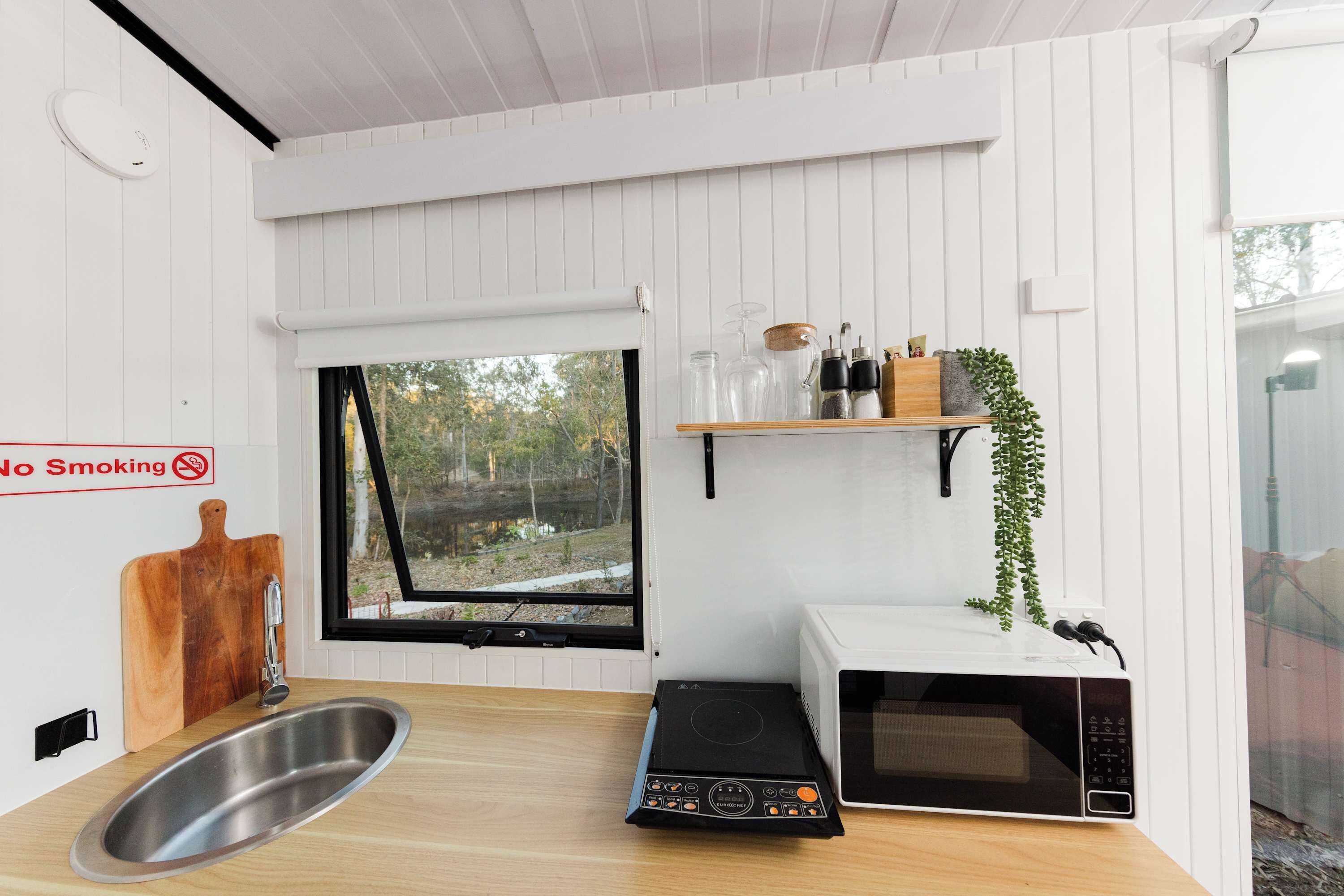 A kitchen with a stainless steel sink, a microwave, a no-smoking sign, and a window with a view of trees.