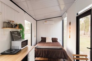 A compact bedroom with a bed, a desk, a microwave, and a small shelf with jars.