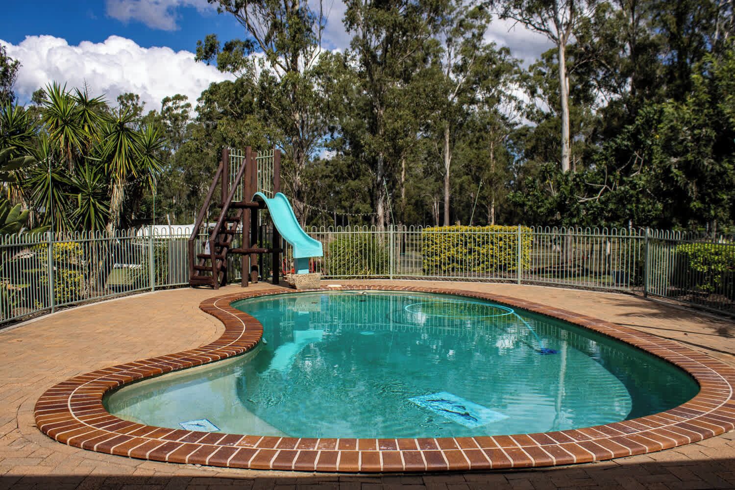 A swimming pool with a slide, surrounded by a brick border and a metal fence.