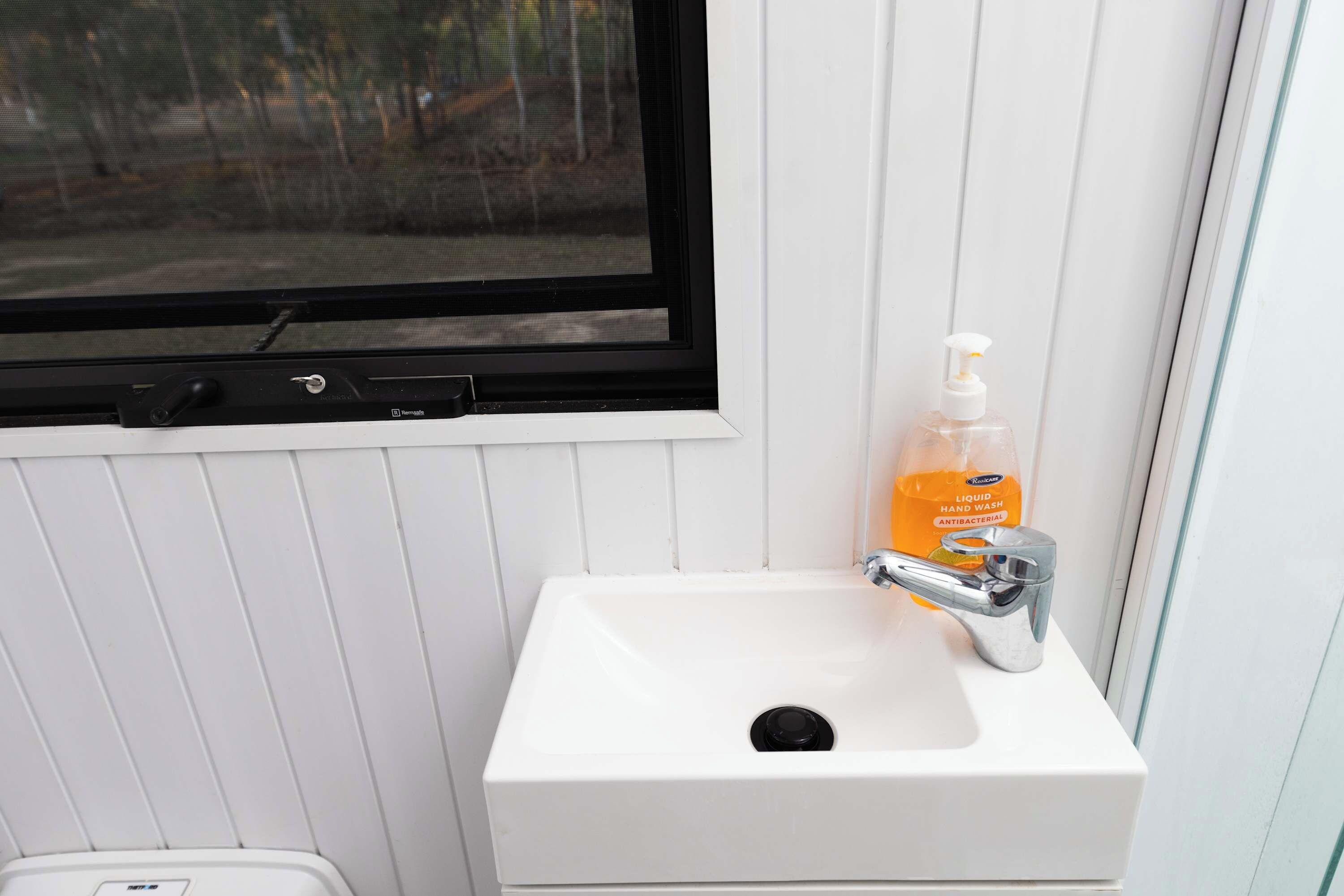 A white bathroom sink with a window above it, a soap dispenser, and a trash bin.
