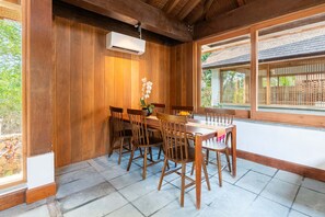 A dining area with a wooden table and chairs, an air conditioning unit, and a view of a traditional building through large windows. - Private Villa with Pool and Garden Views (Nusa Lembongan)