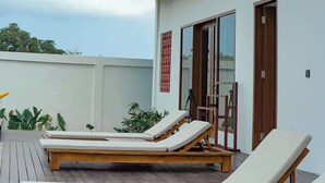 Un edificio moderno de dos pisos con un balcón, una terraza de madera con sillones de descanso y un cielo despejado.