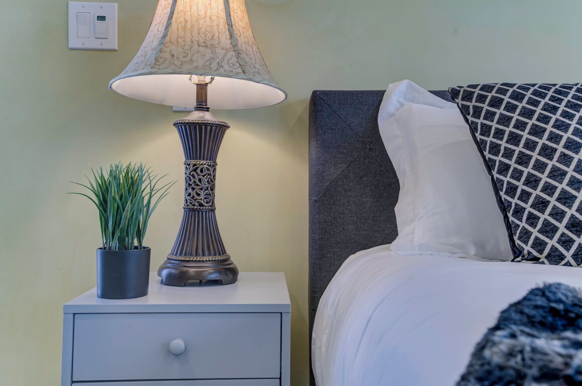 Une table de chevet avec une lampe, une plante en pot et un lit agrémenté d’oreillers à motifs.