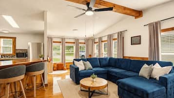 A spacious living area with a blue sectional sofa, a round coffee table, and a dining area with a kitchen in the background.