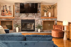 A living room with a stone fireplace, a flat-screen TV mounted above it, and a blue sofa. - NEW! Hot Tub + Sauna, Game Room, Near Red River Gorge (Stanton)