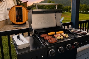 A charcoal grill with multiple cooking zones and a built-in smoker, set on an outdoor deck with a wooden barrel and a view of a green landscape. - NEW! Hot Tub + Sauna, Game Room, Near Red River Gorge (Stanton)