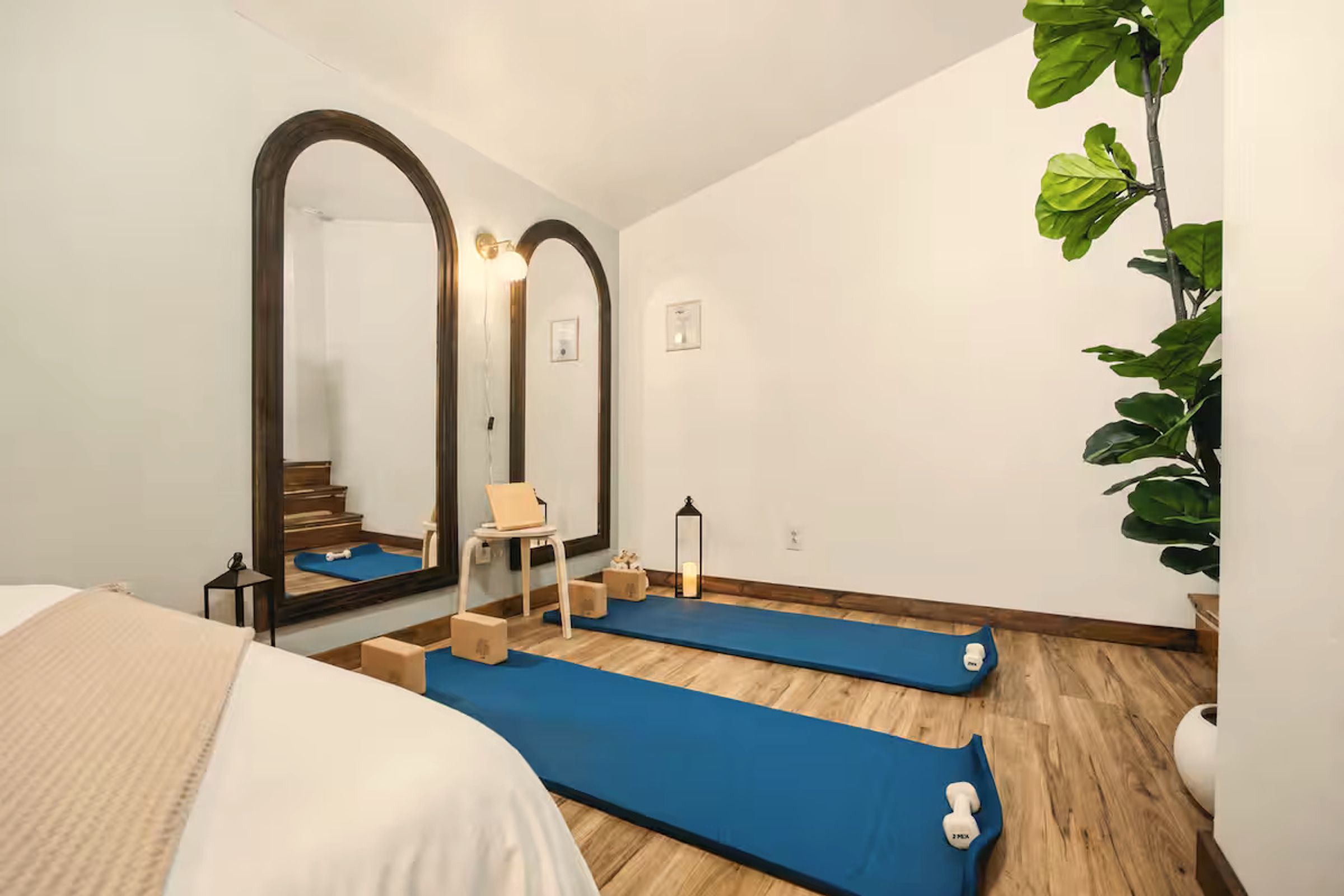 A room with a bed, two blue exercise mats, a chair, a small table, a mirror, and a potted plant.