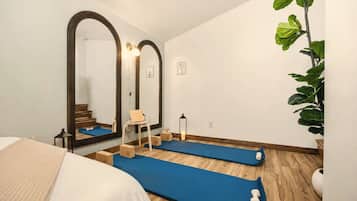 A room with a bed, two blue exercise mats, a chair, a small table, a mirror, and a potted plant.