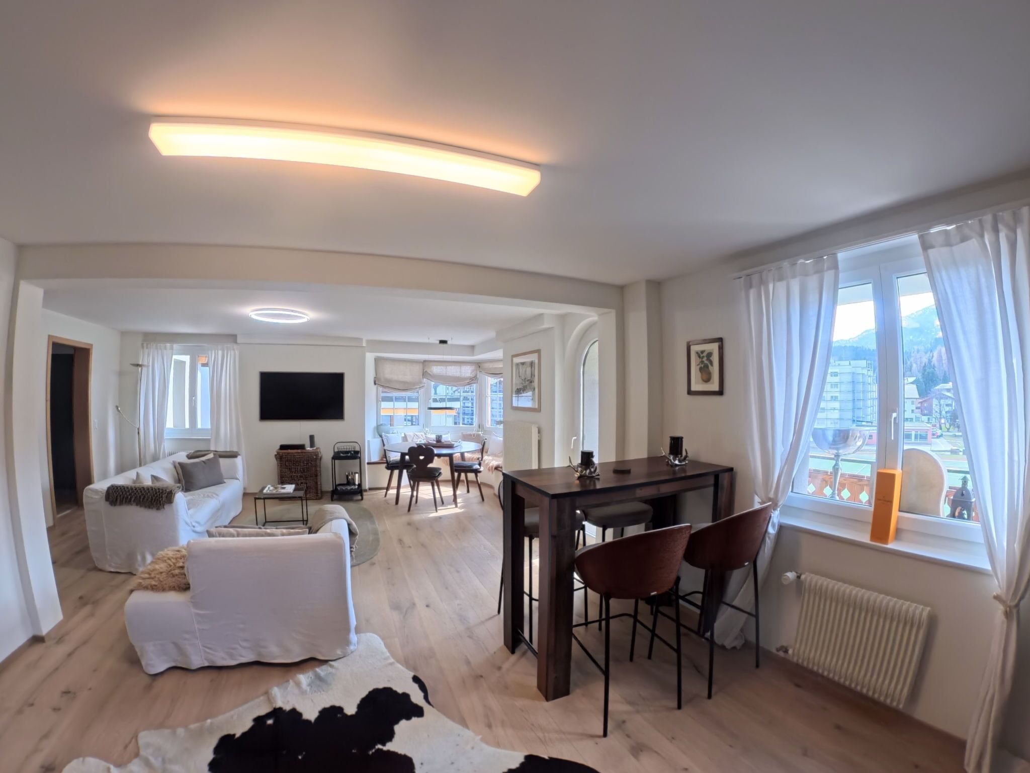 Una sala moderna con un sofá, una mesa de comedor con sillas, un televisor montado en la pared y vistas de la ciudad a través de amplios ventanales.