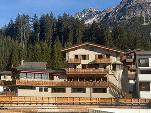 Ein mehrstöckiges Gebäude mit Balkonen, ein Skiabhang im Vordergrund und eine Berglandschaft im Hintergrund.