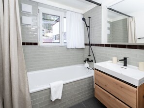 A modern bathroom with a bathtub, a vanity with a mirror, and a shower area. - 1624 Aurora 5.5 by Interhome (Vaz)