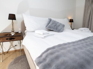 A neatly made bed with white linens, a grey fluffy pillow, and a grey blanket. There are two bedside tables with lamps and a phone. - 1624 Aurora 5.5 by Interhome (Vaz)