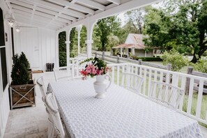 Eine überdachte Veranda mit einem weißen Tisch und Stühlen, einem Blumenstrauß und Blick auf ein Haus und Bäume.