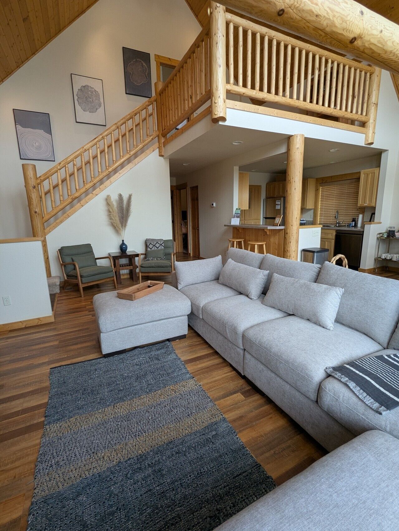 Un vaste espace de vie comprenant un grand canapé d’angle gris, un escalier en bois et une cuisine en arrière-plan.