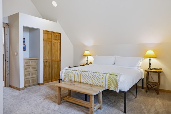 A bedroom with a bed, bedside tables, a bench, a wardrobe, and a door leading to another room.