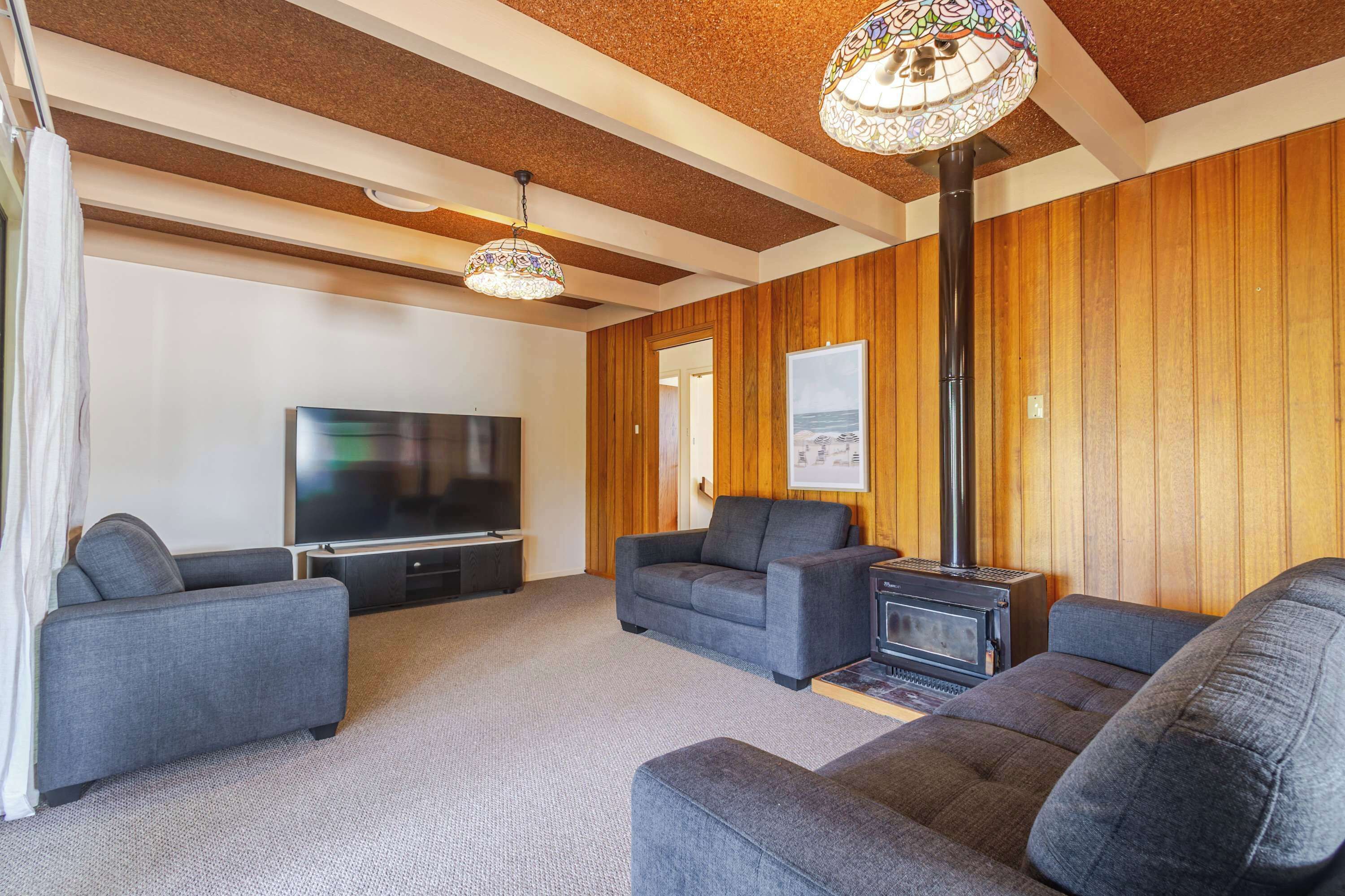 A living room with a flat-screen TV, two grey sofas, a wood-paneled wall, a fireplace, and a ceiling with exposed beams.