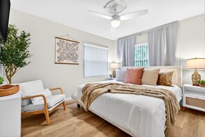 Una camera da letto con un letto, comodini, una sedia, una ventola a soffitto, una finestra con tende e un'opera d'arte appesa al muro.