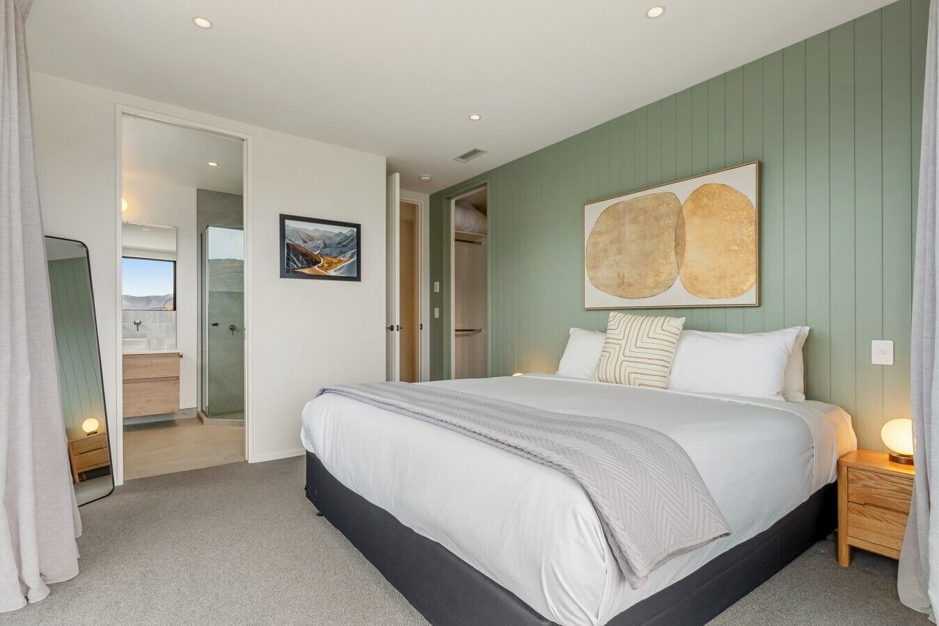 A bedroom with a large bed, a nightstand, a wall-mounted artwork, and a bathroom visible through an open door.