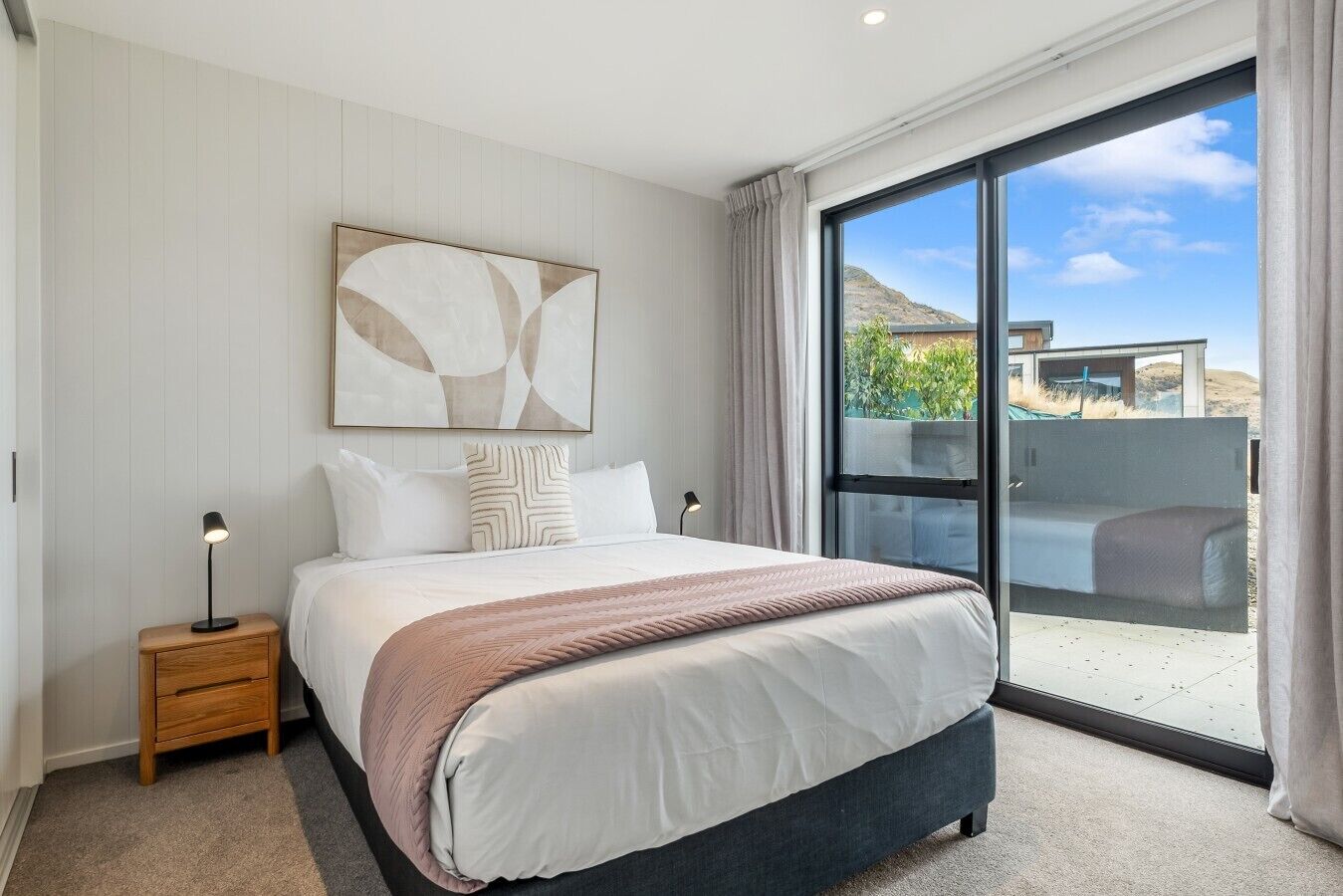 A bedroom with a large bed, a nightstand, a wall-mounted artwork, and a view through a sliding glass door.