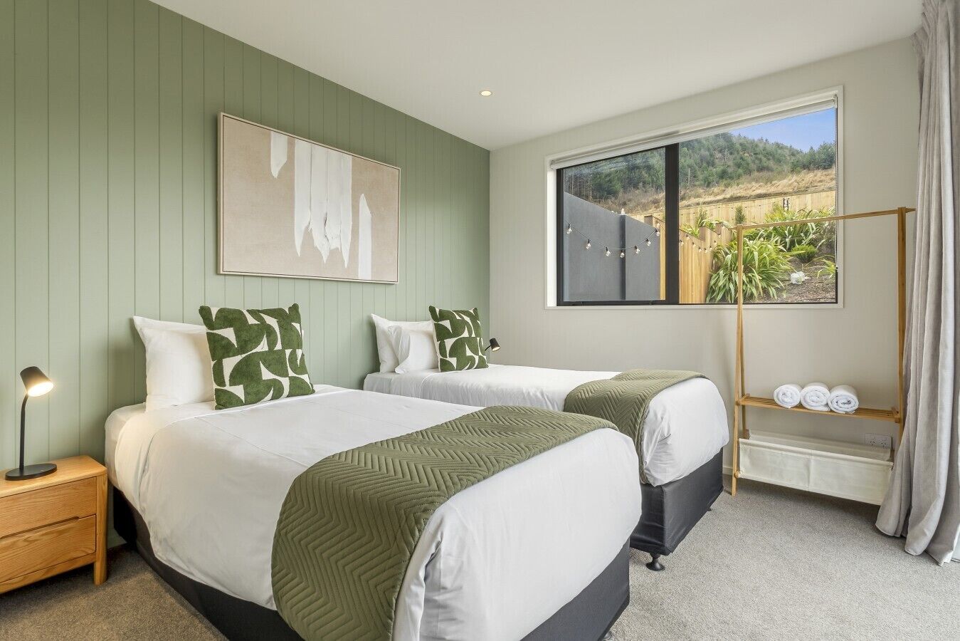 A hotel room with two beds, a nightstand, a lamp, a window with a view, and a shelf with towels.