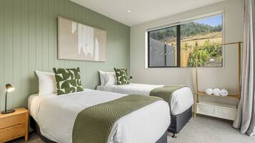 A hotel room with two beds, a nightstand, a lamp, a window with a view, and a shelf with towels.