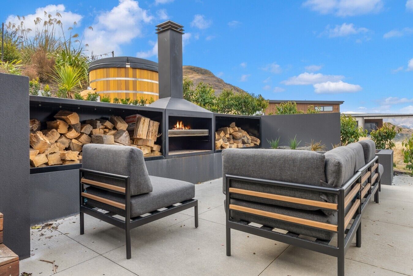 An outdoor seating area with a wood-burning stove, a cylindrical structure, and a view of a hilly landscape.