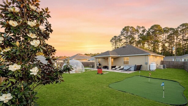 Un patio trasero residencial con una casa, una zona de juegos cubierta, un campo de golfito y un árbol con flores blancas.