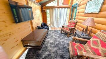 L’intérieur d’une cabane en rondins est agrémenté d’un téléviseur à écran plat fixé au mur, d’un banc en bois, de deux fauteuils à motifs et d’une petite table avec une lampe.